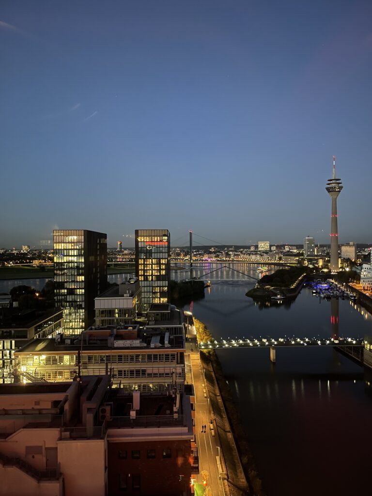 Nachtaufnahme des Düsseldorfer MedienHafens mit beleuchteten Gebäuden, Rhein, Brücken und dem Rheinturm im Hintergrund.